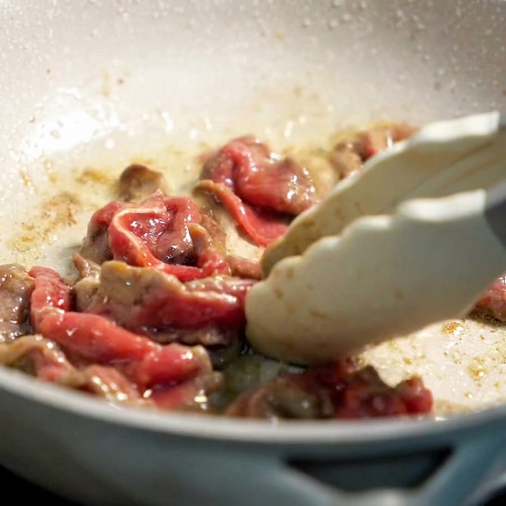 Tranches de bœuf partiellement cuites sautées dans une poêle légèrement huilée avec des pinces en silicone.