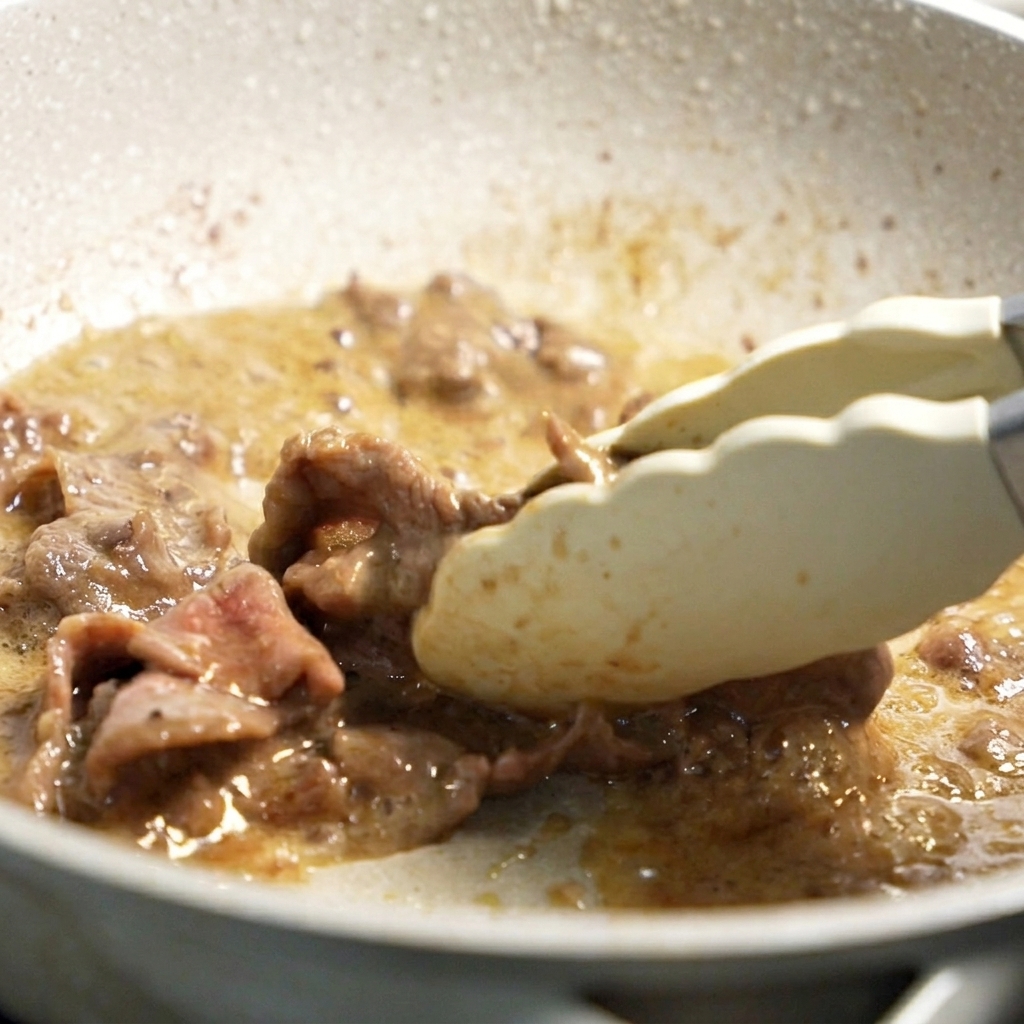 Pinces mélangeant des tranches de bœuf dorées dans une poêle chaude avec de lhuile de cuisson.
