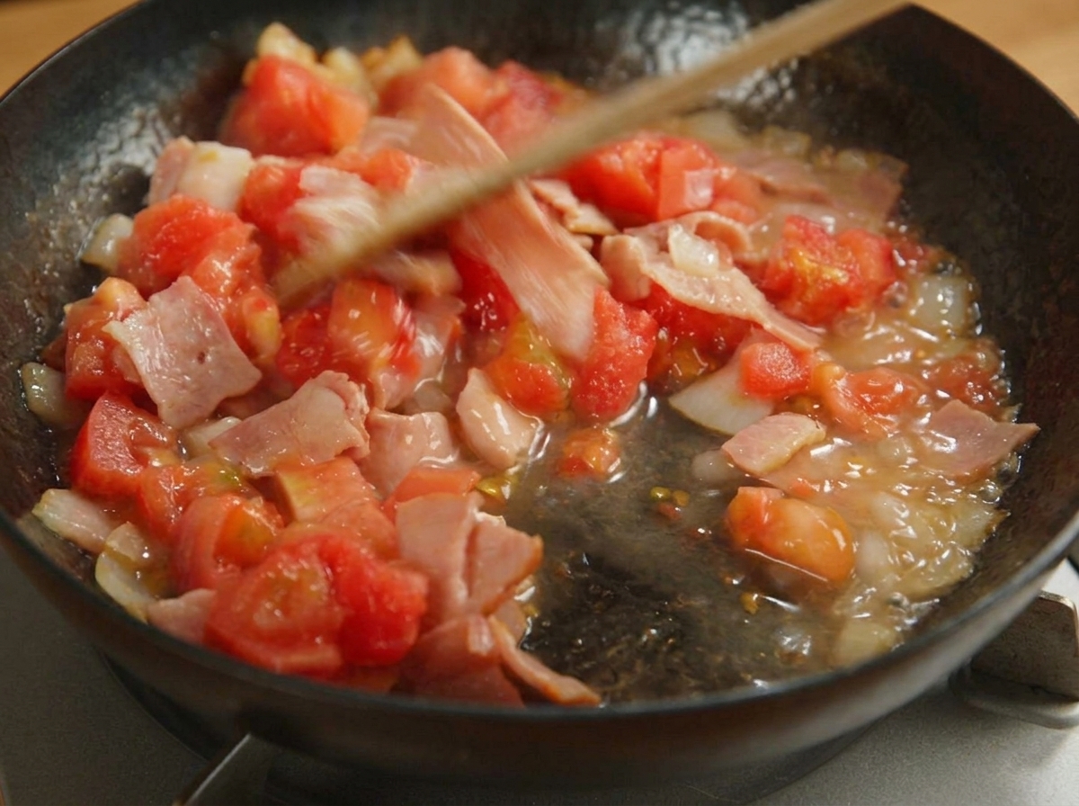 Spatule en bois remuant les tomates, le bacon et les oignons dans une poêle.