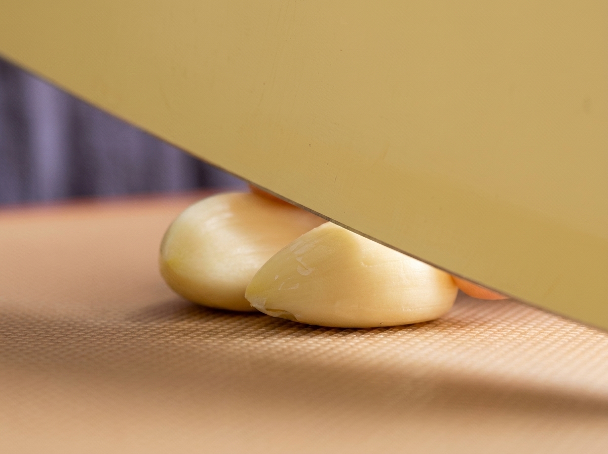 Un grand couteau de cuisine posé sur deux gousses dail pelées sur une planche à découper marron texturée, prêtes à être hachées.