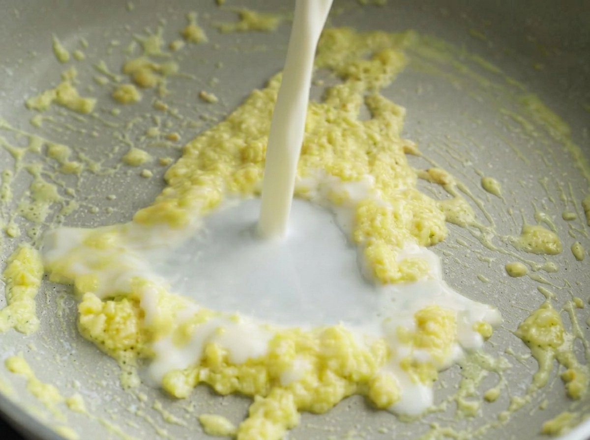 Un filet de lait blanc versé dans une poêle contenant un mélange grésillant de beurre jaune et dail haché.
