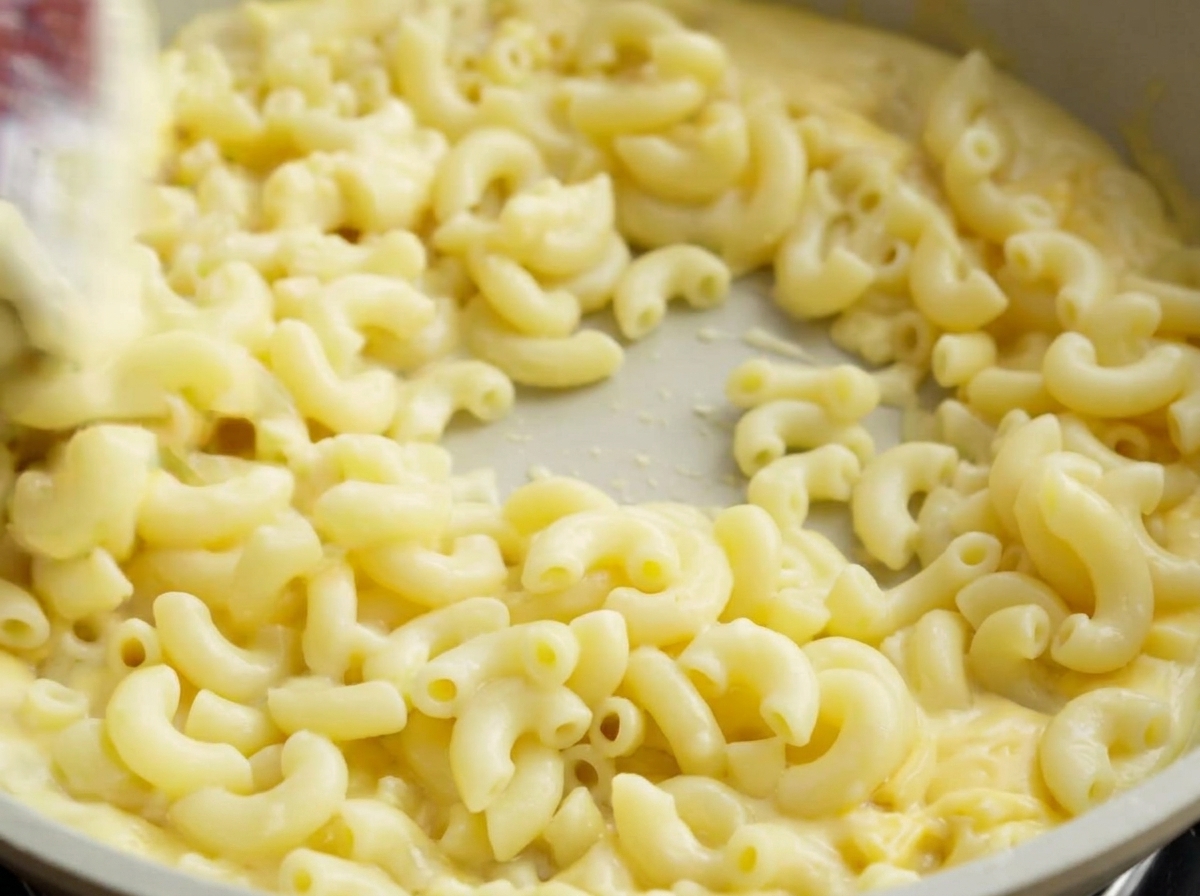Une spatule remuant des macaronis cuits dans une poêle de couleur claire, enrobant parfaitement les pâtes de sauce au fromage crémeuse.