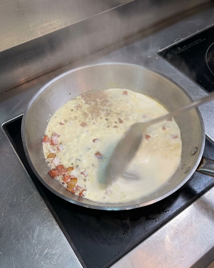 Poêle en inox avec la crème frémissante et le bacon, remuée à la cuillère.