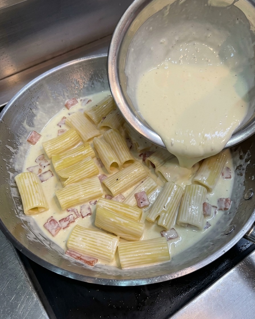 Mélange jaune dœuf, crème et fromage versé sur les rigatoni et le bacon dans la poêle.