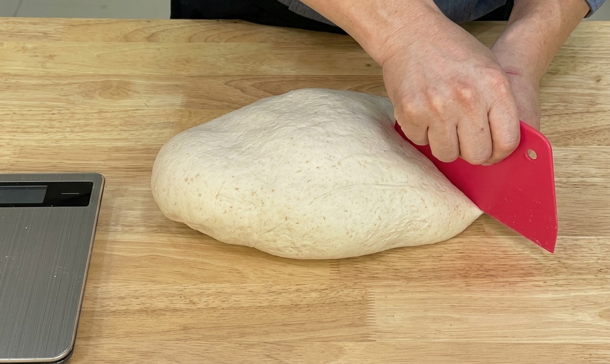 Chef utilisant un coupe-pâte rouge pour diviser la pâte lisse sur un plan de travail en bois.