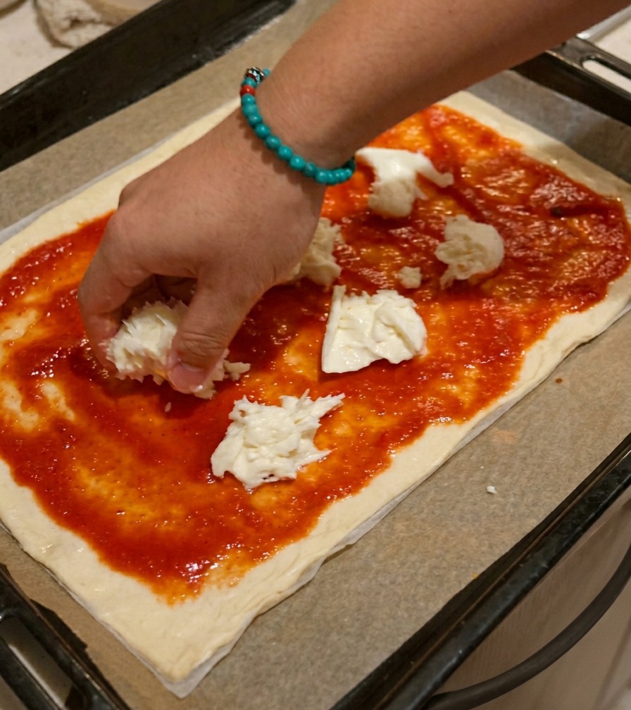 Une main déposant des morceaux de mozzarella blanche déchirés à la main sur la sauce tomate.