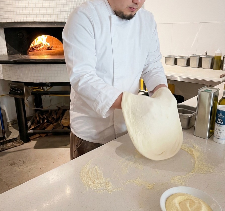 Un chef presse et étire un pâton de pâte à pizza rond sur un plan de travail blanc fariné.