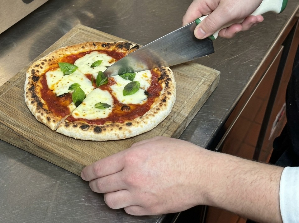 Mains utilisant un grand couteau pour couper une pizza Margherita en parts sur une planche à découper.