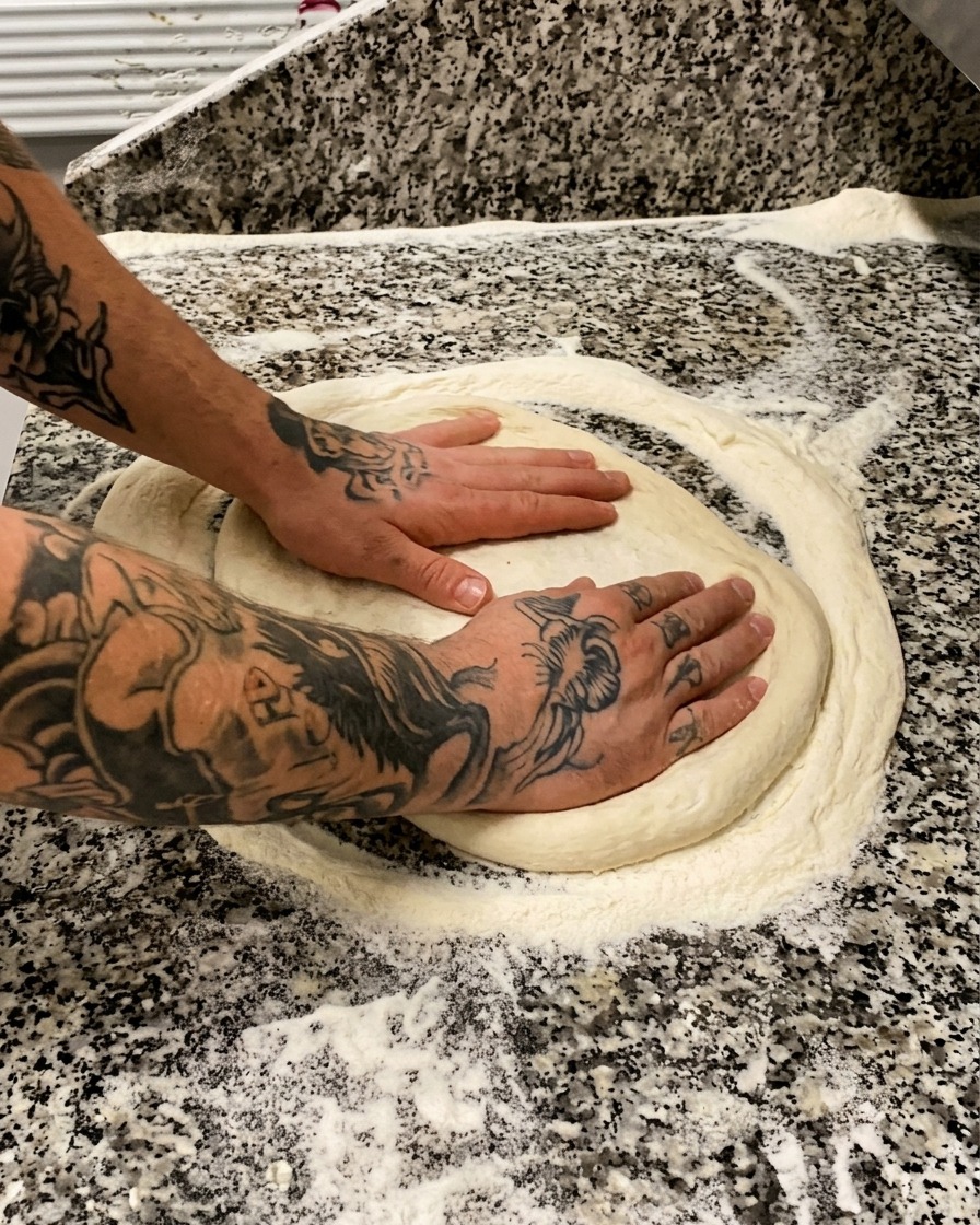 Des mains tatouées pressant et étirant délicatement une pâte à pizza ronde sur une surface en marbre farinée.