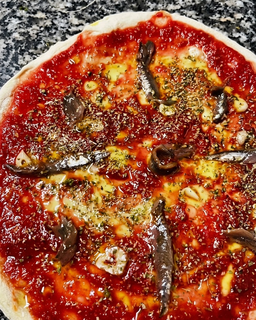 Vue rapprochée de dessus dune pizza crue garnie de sauce tomate, dhuile dolive, dorigan et de filets danchois marinés dispersés.