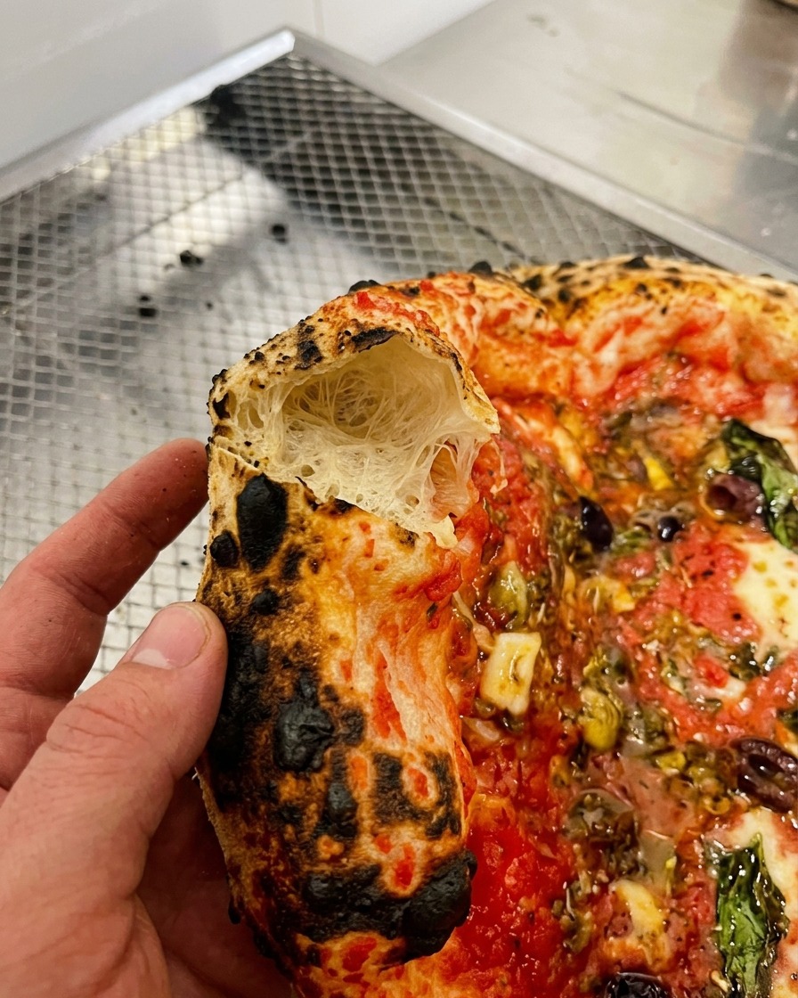 Gros plan dune main tenant un morceau de croûte de pizza montrant une structure intérieure très aérée et alvéolée.