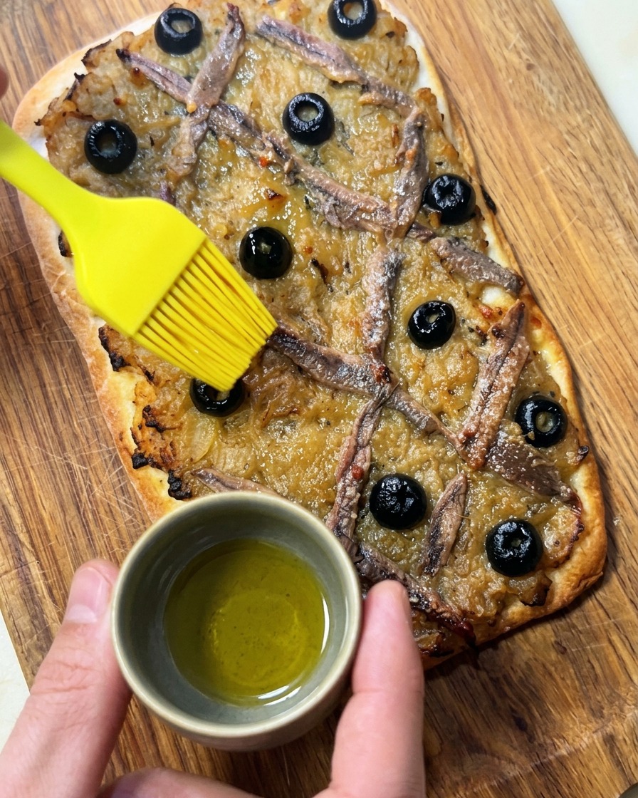 Une main utilisant un pinceau en silicone pour appliquer de lhuile dolive sur la pissaladière cuite.