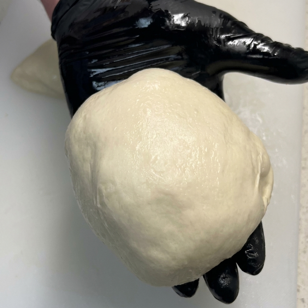 Une boule de pâte à pizza parfaitement lisse reposant dans la paume dune main gantée.