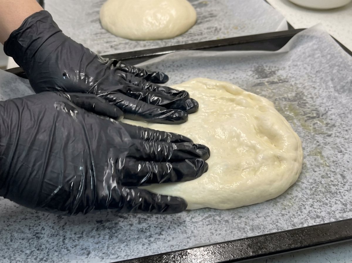 Des mains gantées pressant et étirant une pâte souple sur une plaque de cuisson tapissée de papier sulfurisé.