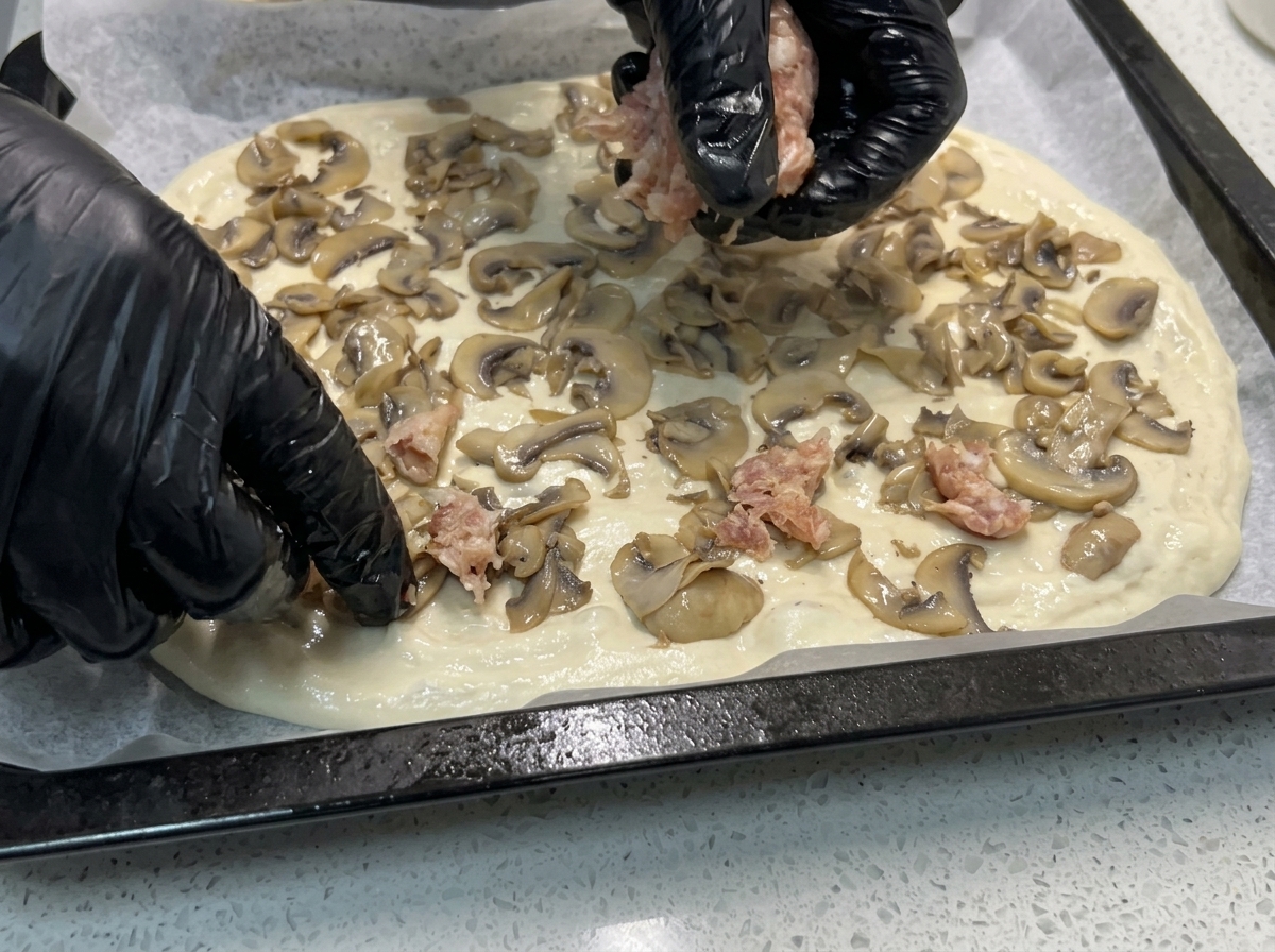 Mains déposant des morceaux de chair à saucisse crue sur la pâte déjà garnie de champignons.