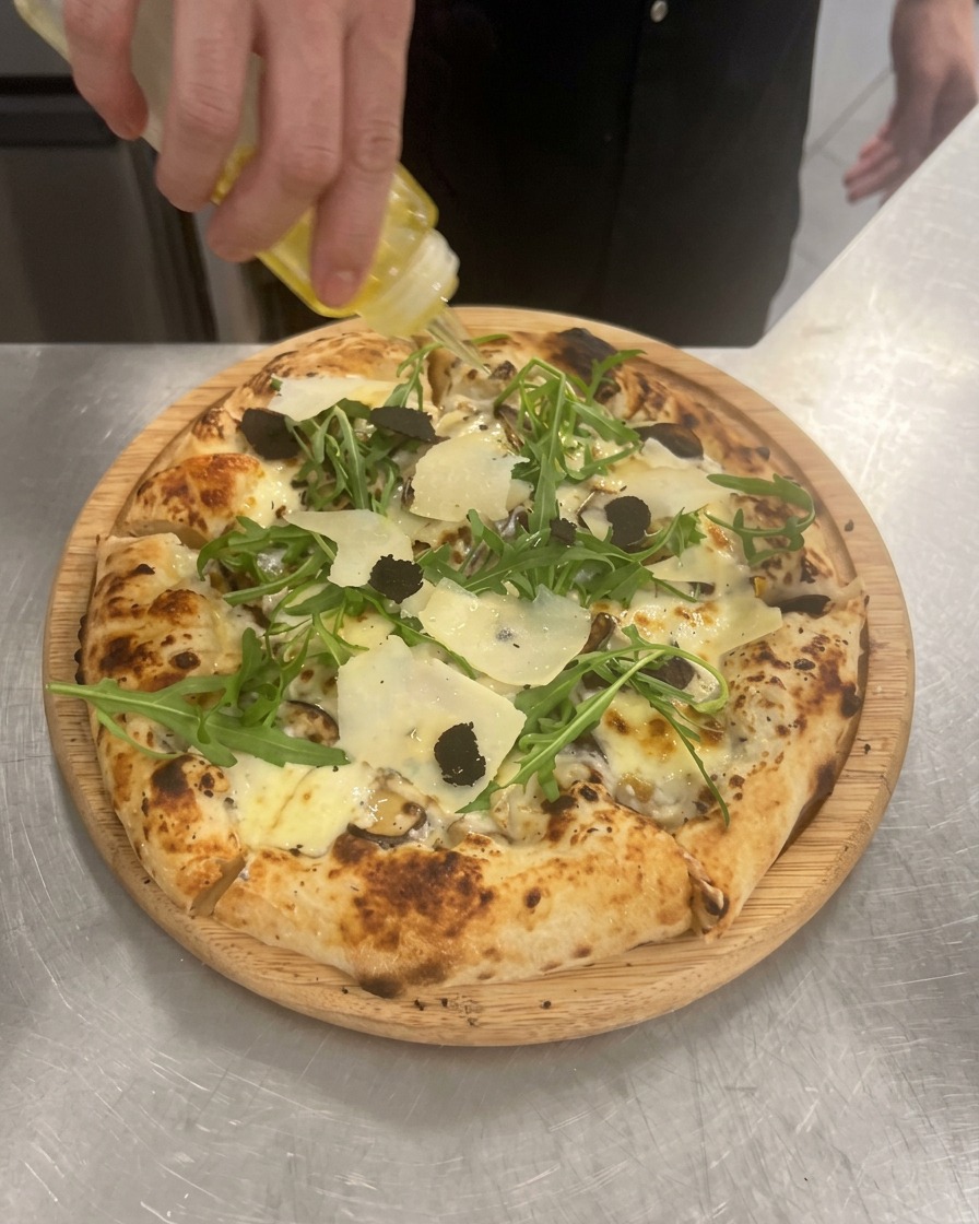 Une main pressant une bouteille transparente pour verser un filet dhuile de truffe dorée sur la pizza.