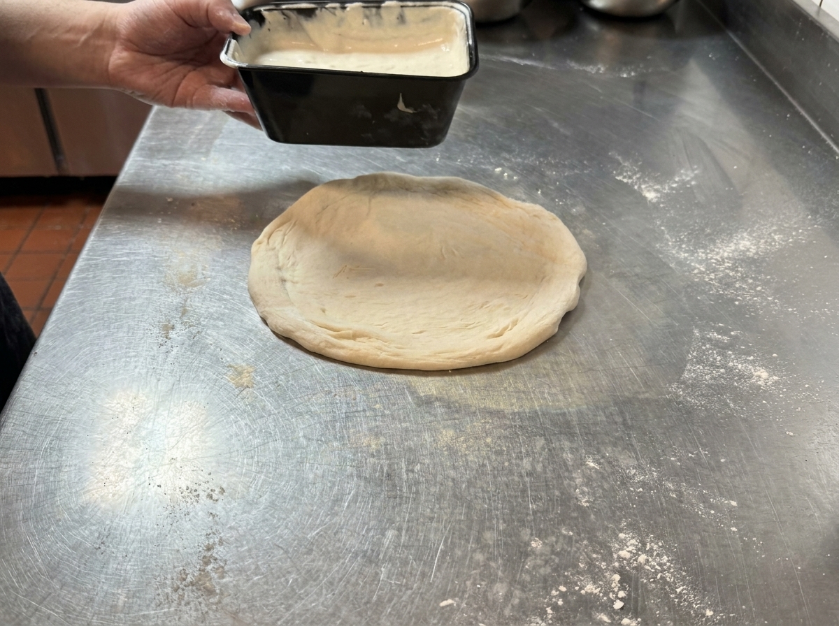 Une main versant de la sauce blanche crémeuse à la truffe sur une pâte à pizza crue et étalée, reposant sur une table en inox.