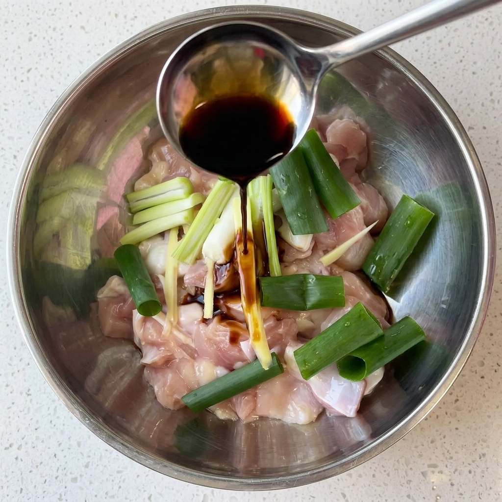 Morceaux de poulet cru dans un bol en métal avec de la sauce soja, des oignons verts et du gingembre.