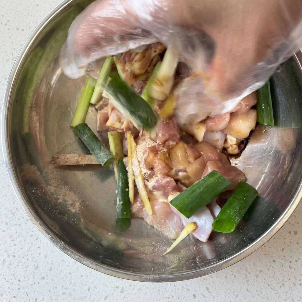 Une main gantée mélangeant le poulet avec les oignons verts, le gingembre et la marinade dans un bol.