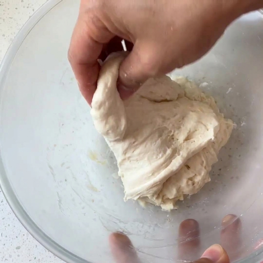 Une main attrapant et pliant une boule de pâte humide et collante dans un bol en verre.