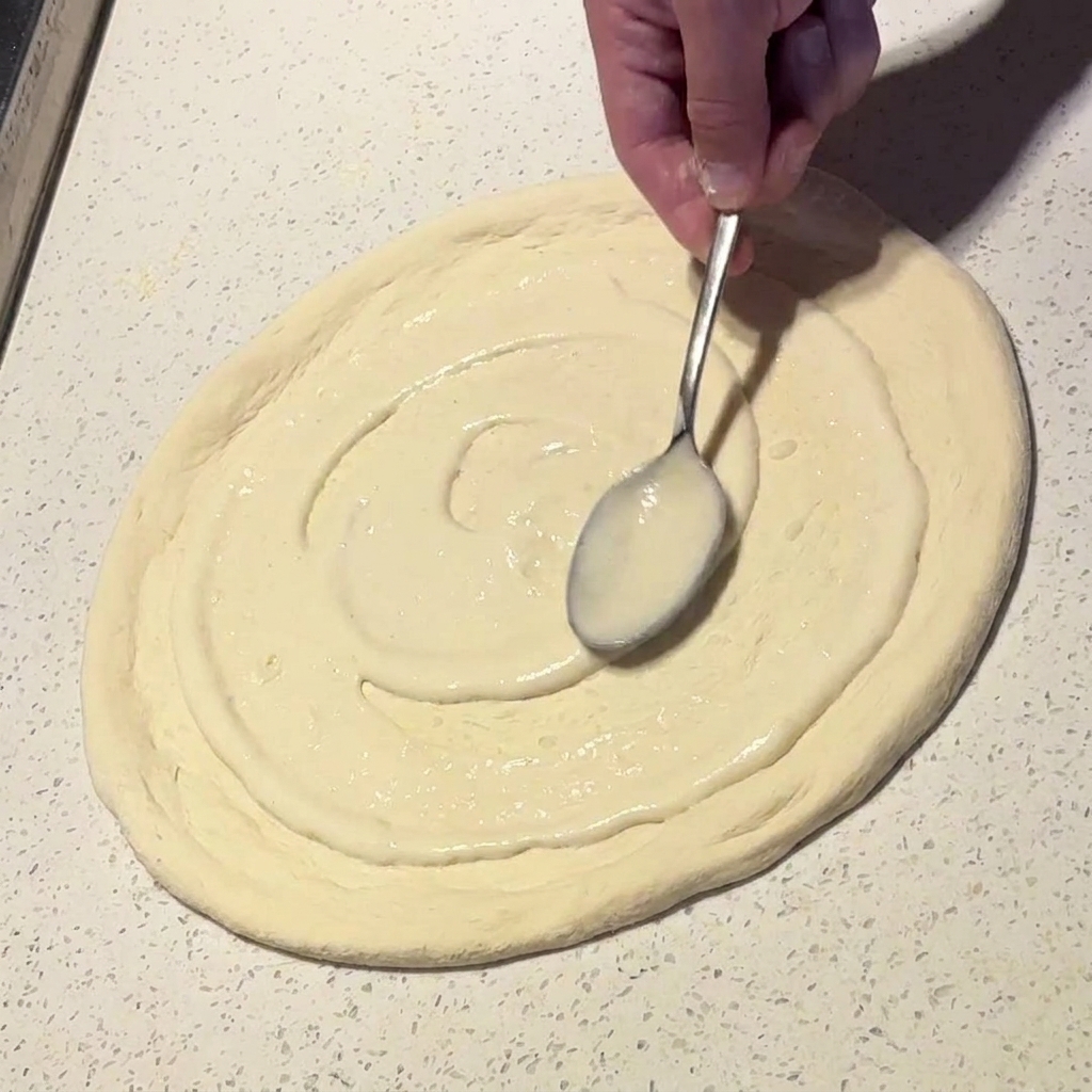 Une cuillère métallique étalant une sauce blanche crémeuse en mouvement circulaire sur une base de pizza.
