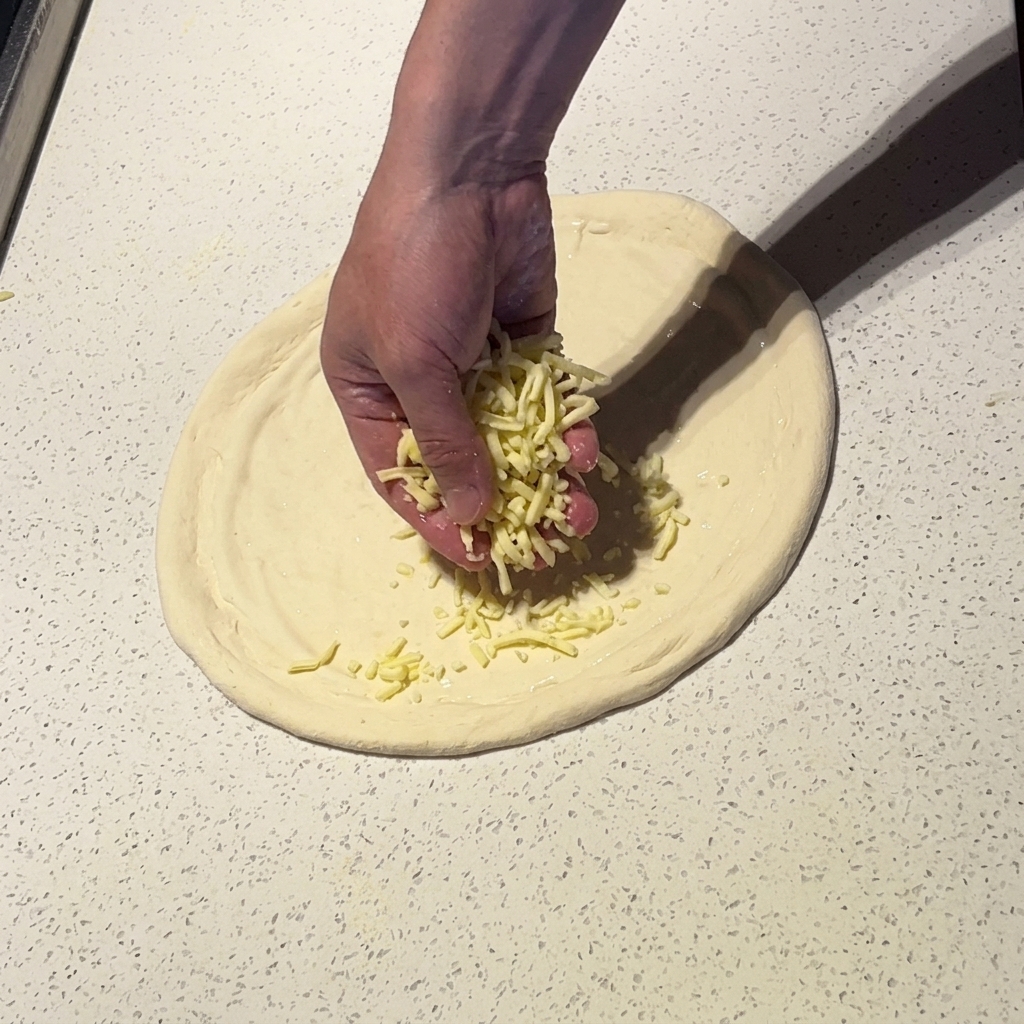 Une main saupoudrant de la mozzarella râpée sur une pâte à pizza nappée de sauce blanche.
