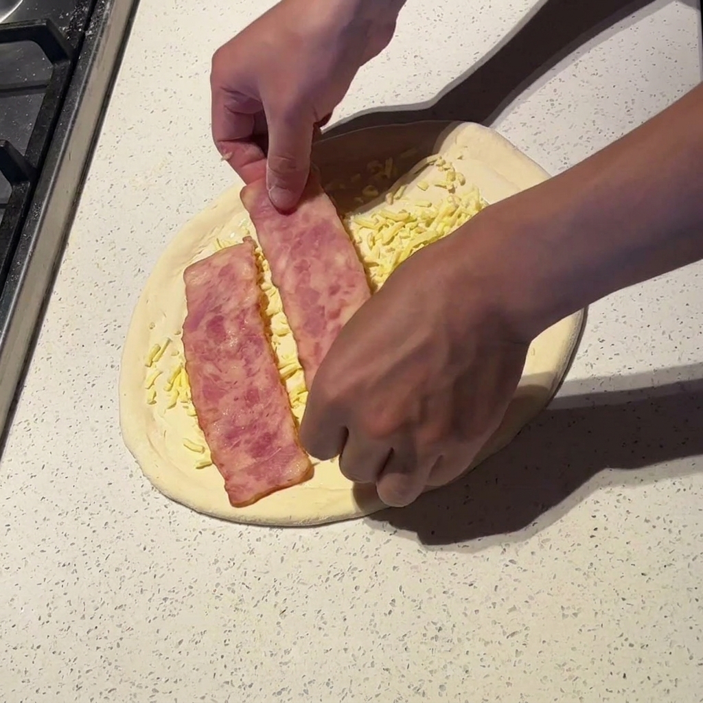 Mains disposant de larges tranches de bacon légèrement cuites sur une pizza garnie de sauce et de fromage.