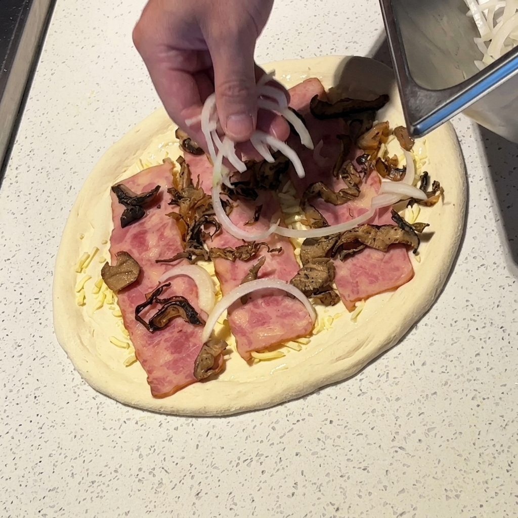 Une main parsemant de fines tranches doignon blanc sur une pizza garnie de bacon et de champignons.