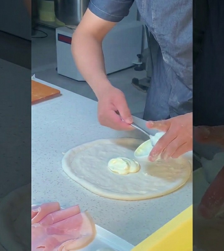 Un chef utilise le dos dune cuillère pour étaler uniformément une sauce blanche riche sur la pâte étirée en un mouvement circulaire.