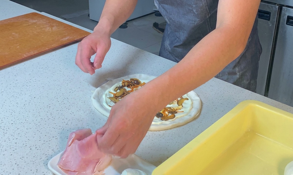 Mains dun chef déchirant des tranches de jambon rose et les répartissant sur une pizza garnie de sauce et de champignons.