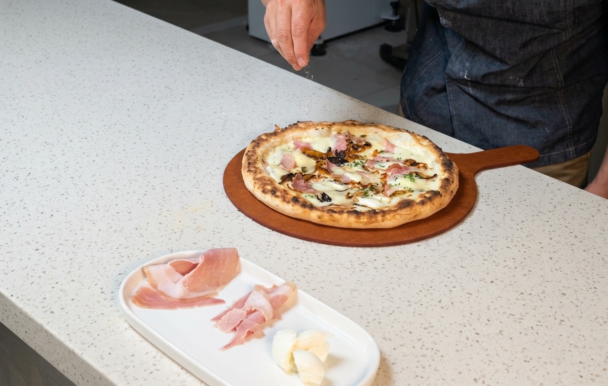 La main dun chef saupoudrant du fromage râpé sur une pizza au jambon et champignons sortant du four.