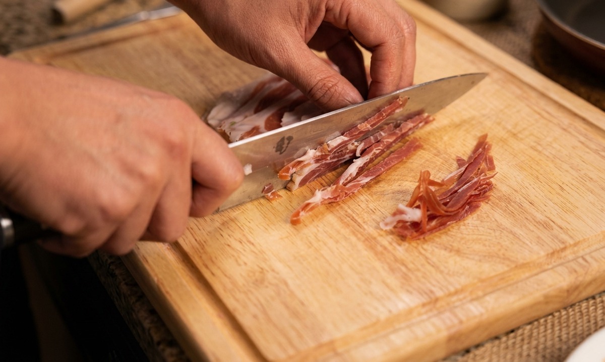 Mains utilisant un couteau de chef pour couper de fines lanières de jambon cru.
