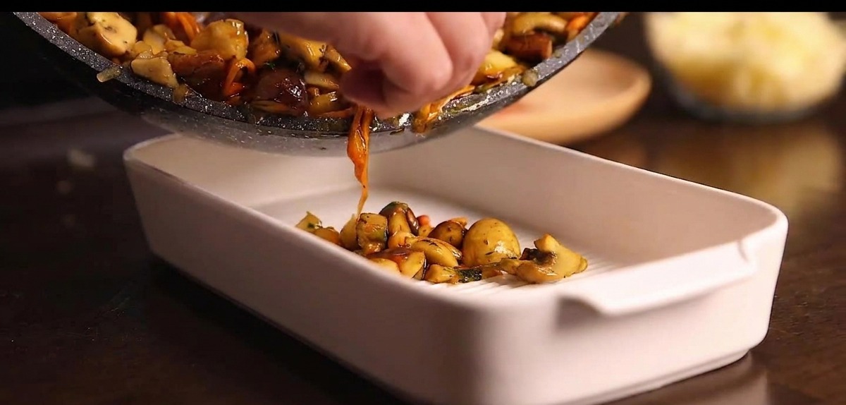 Poêle versant le mélange de champignons cuits dans un plat à gratin en céramique blanche.