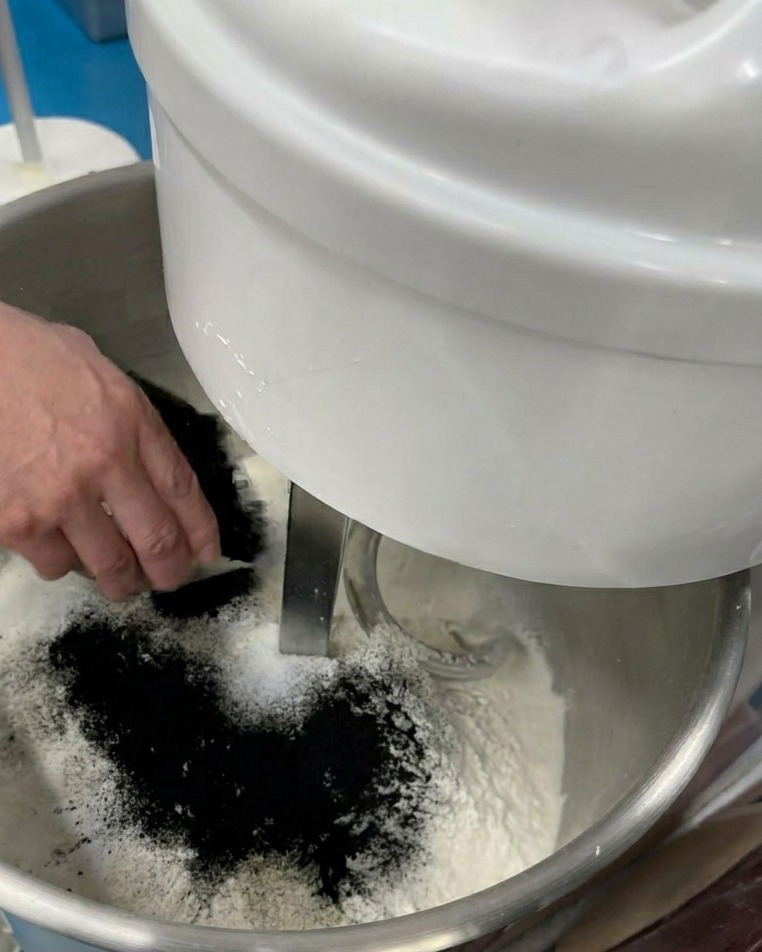 Une main versant une fine poudre noire dans un bol mélangeur rempli de farine et dingrédients secs.