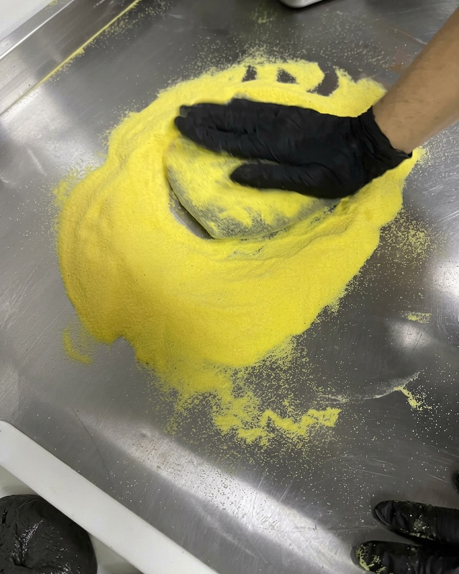 Mains gantées étirant une pâte noire en cercle sur une surface en inox saupoudrée de farine jaune.