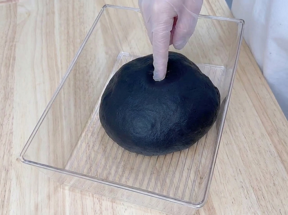 Un doigt ganté enfoncé au centre dune boule de pâte noire ayant bien levé dans son bac.