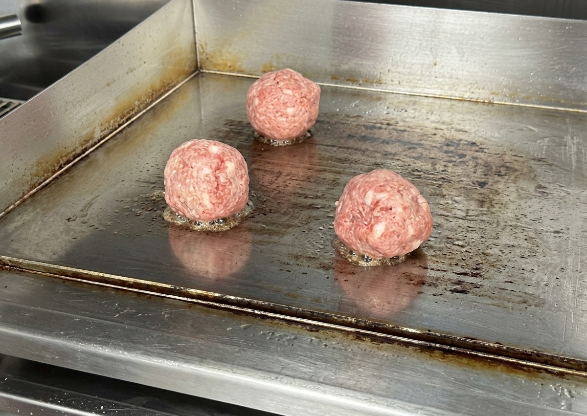 Trois boules de bœuf crues posées sur une plaque de cuisson en acier inoxydable chaude avec un léger grésillement.