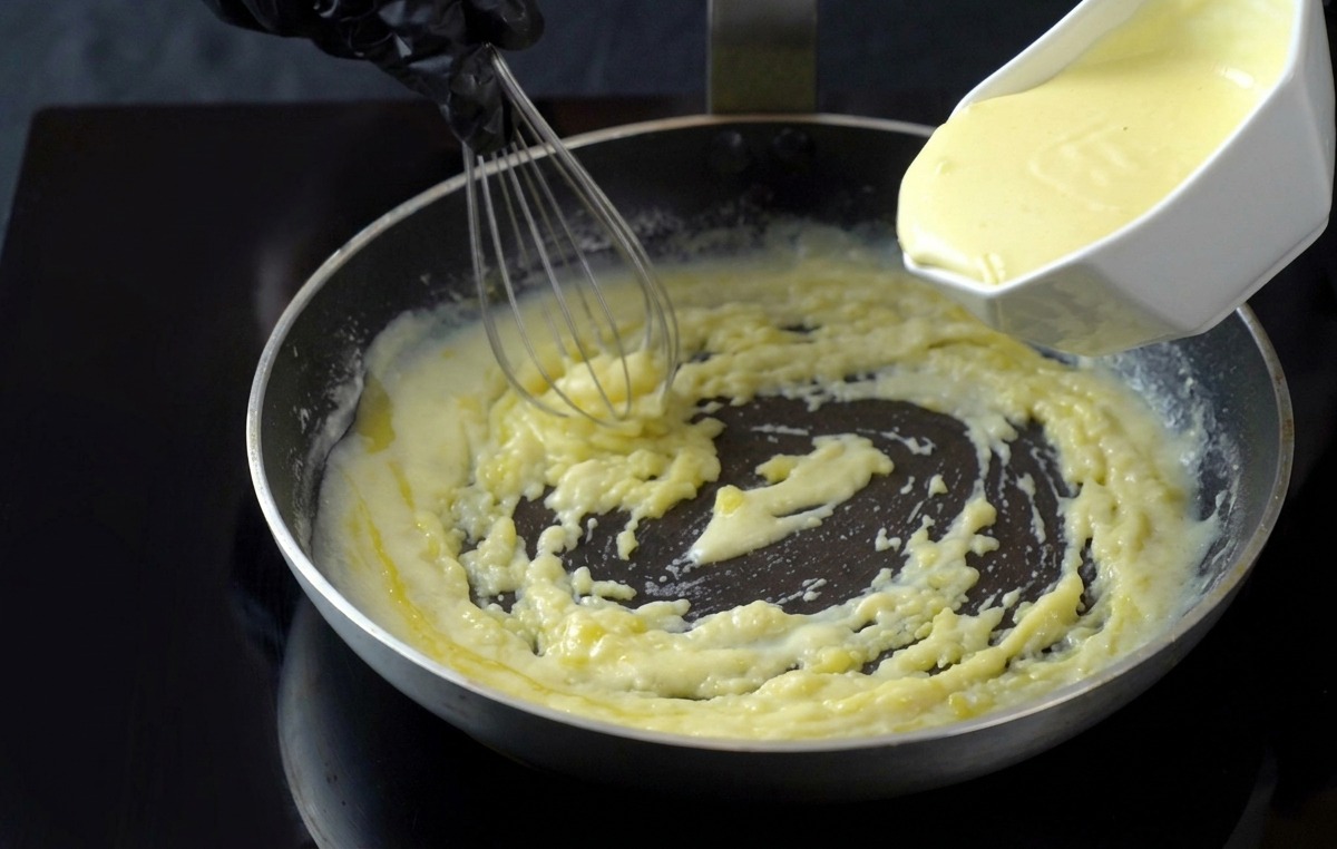 Verser de la crème liquide dun récipient blanc dans une sauce blanche frémissante dans une poêle, en mélangeant avec un fouet.