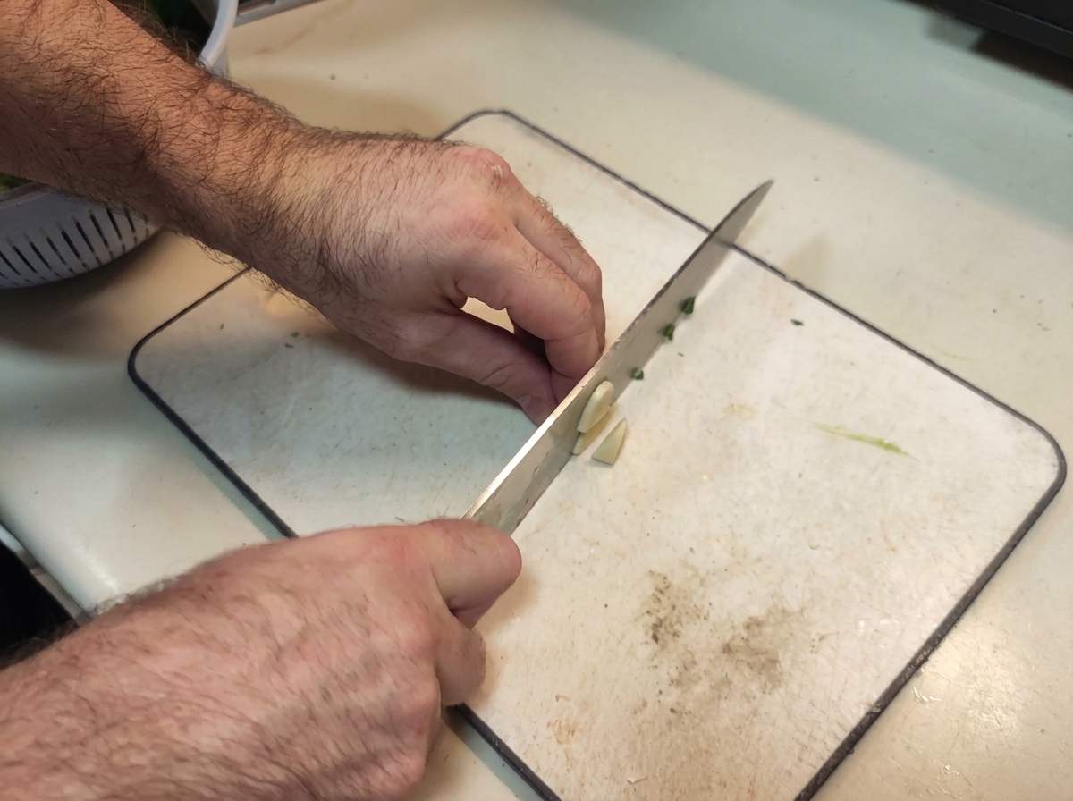 Mains utilisant un couteau de chef pour émincer finement des gousses dail sur une planche blanche.