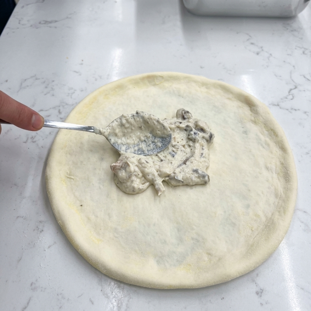 Une main utilisant le dos dune cuillère en métal pour étaler une sauce blanche aux champignons sur la pâte crue.