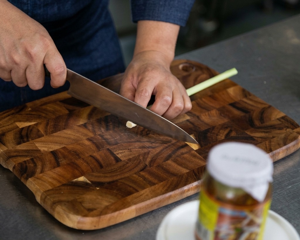 Mains utilisant un couteau aiguisé pour hacher finement des bâtons de citronnelle fraîche sur une planche en bois.