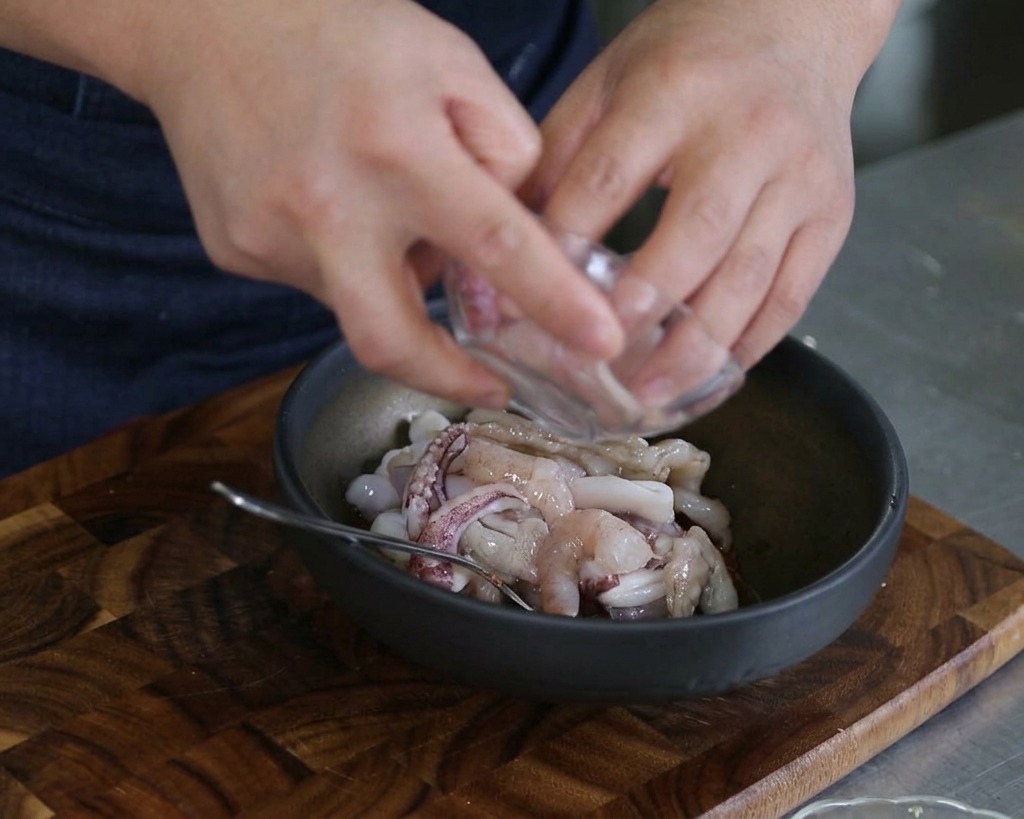 Mains versant des morceaux de calmar et des crevettes décortiquées dans un bol de mélange avec les autres ingrédients.