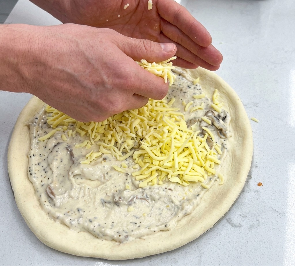 Deux mains saupoudrant des poignées de mozzarella râpée sur une base de pizza nappée de sauce.
