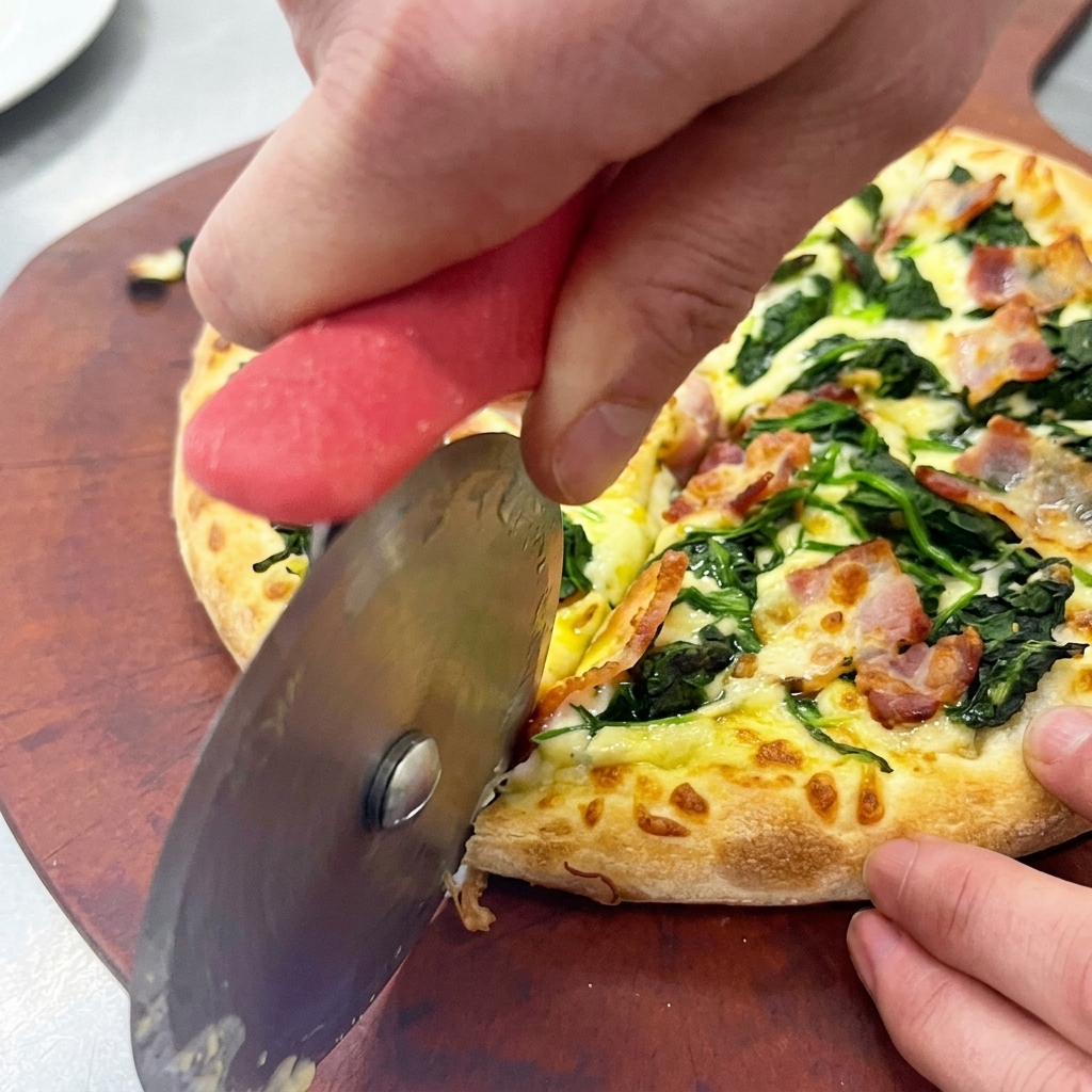 Mains utilisant une roulette à pizza pour découper une pizza aux épinards et au bacon sur une planche en bois.