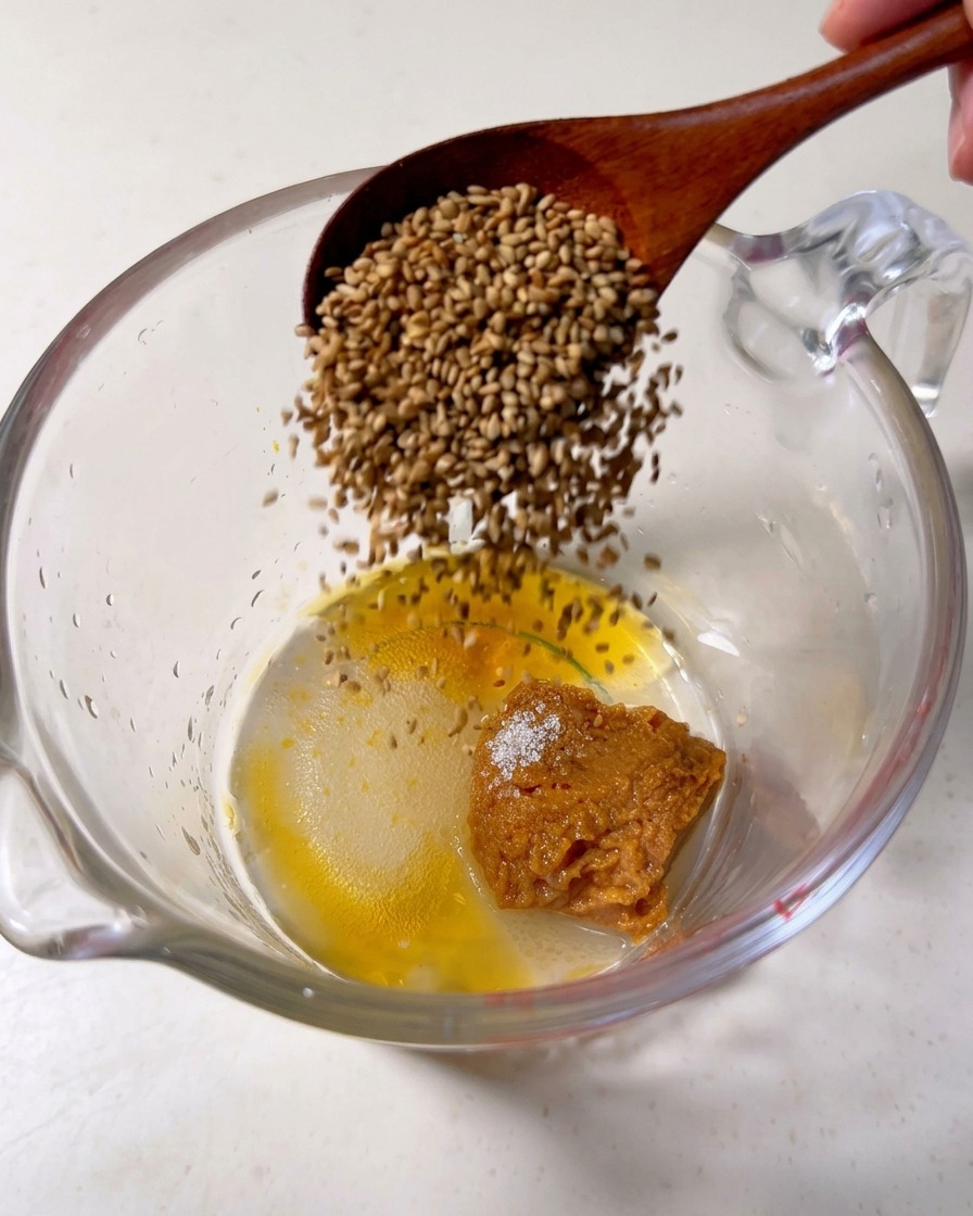 Une cuillère en bois versant des graines de sésame dans un récipient contenant du miso, du sucre et de lhuile de sésame.