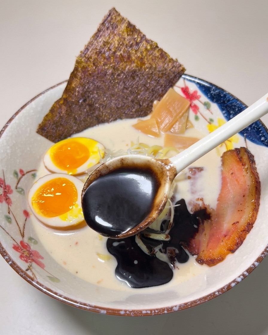 Une cuillère versant de lhuile dail noir épaisse et sombre sur le ramen terminé.