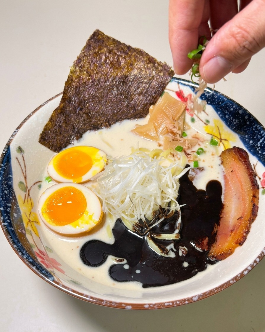 Une main saupoudrant des oignons verts hachés sur un bol de ramen magnifiquement dressé.