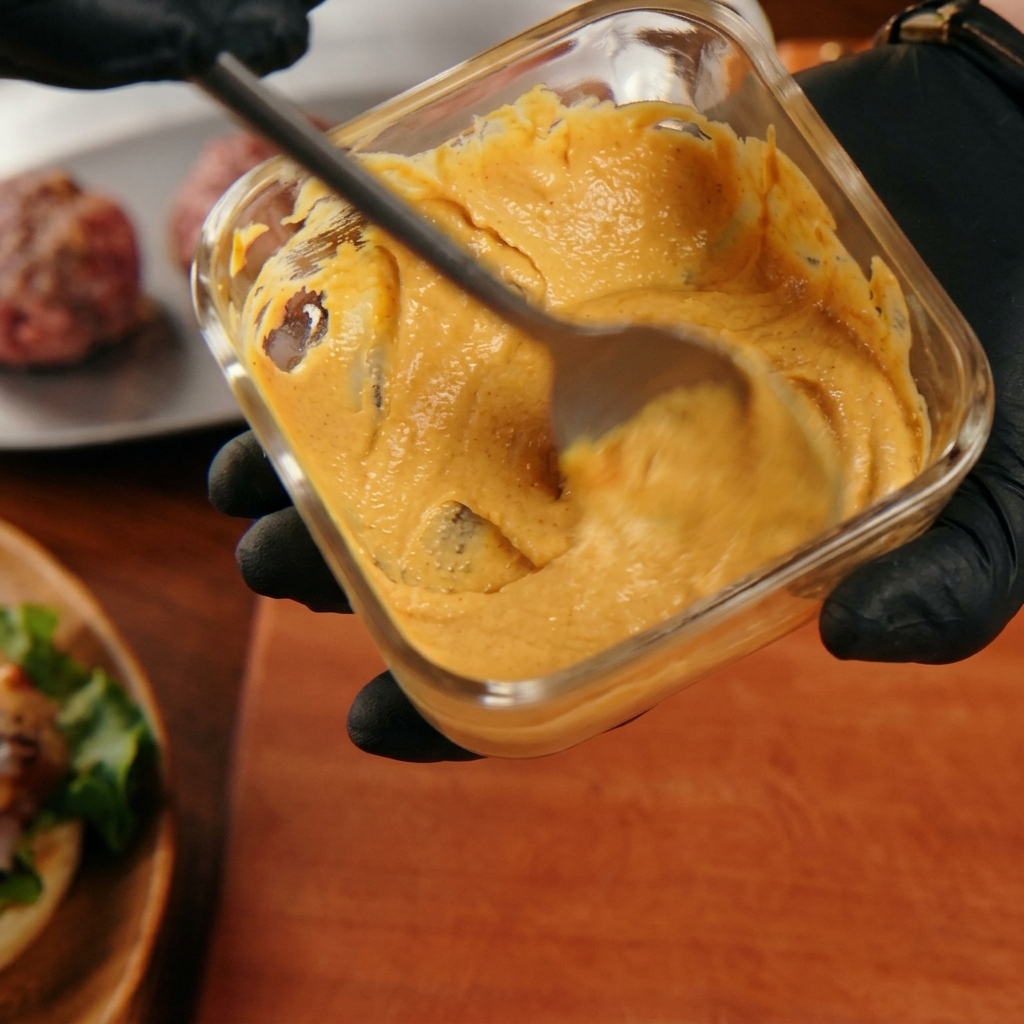 Une main mélangeant une sauce burger orange crémeuse dans un récipient en verre.