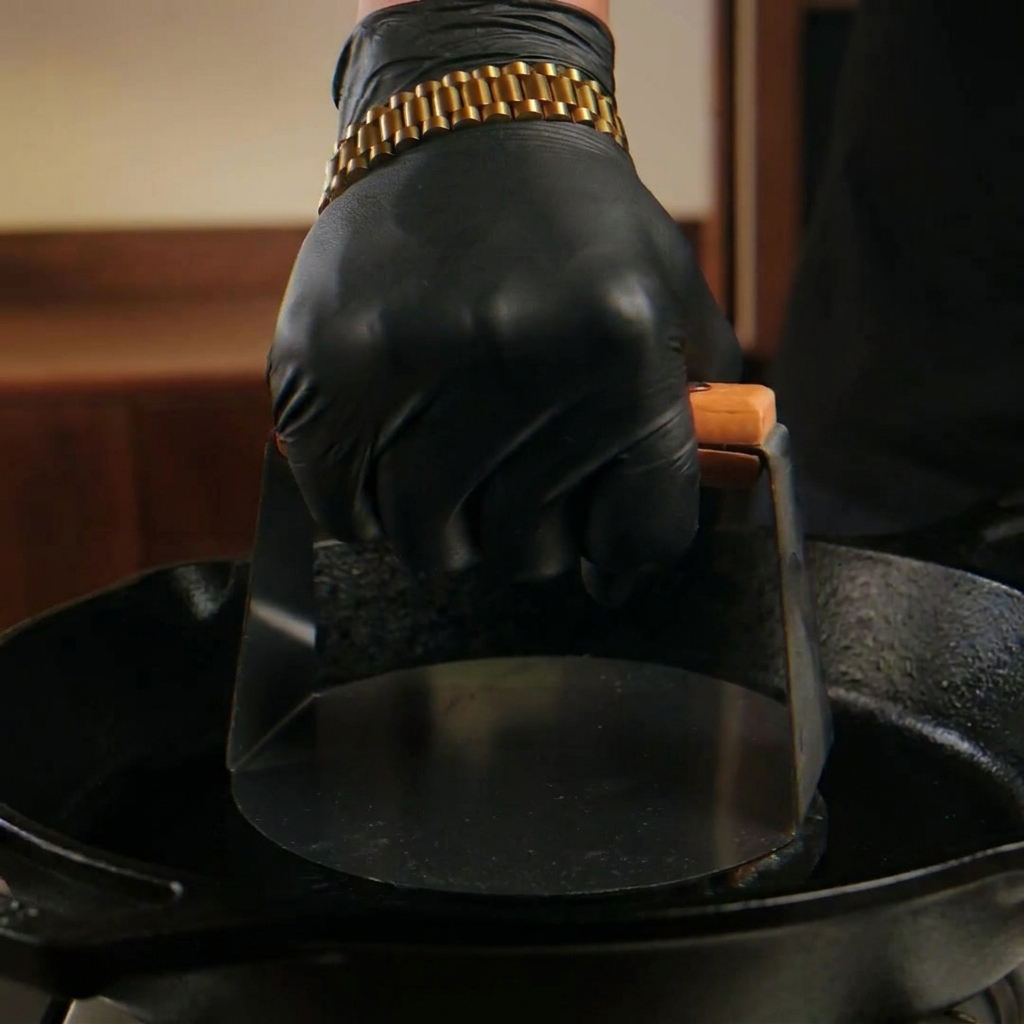 Une main pressant fermement une boulette de bœuf sur une poêle en fonte fumante avec une presse métallique.