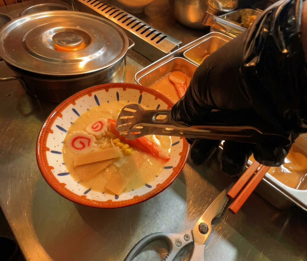 Une main déposant des tranches de narutomaki (pâte de poisson) dans un bol de ramen fumant.